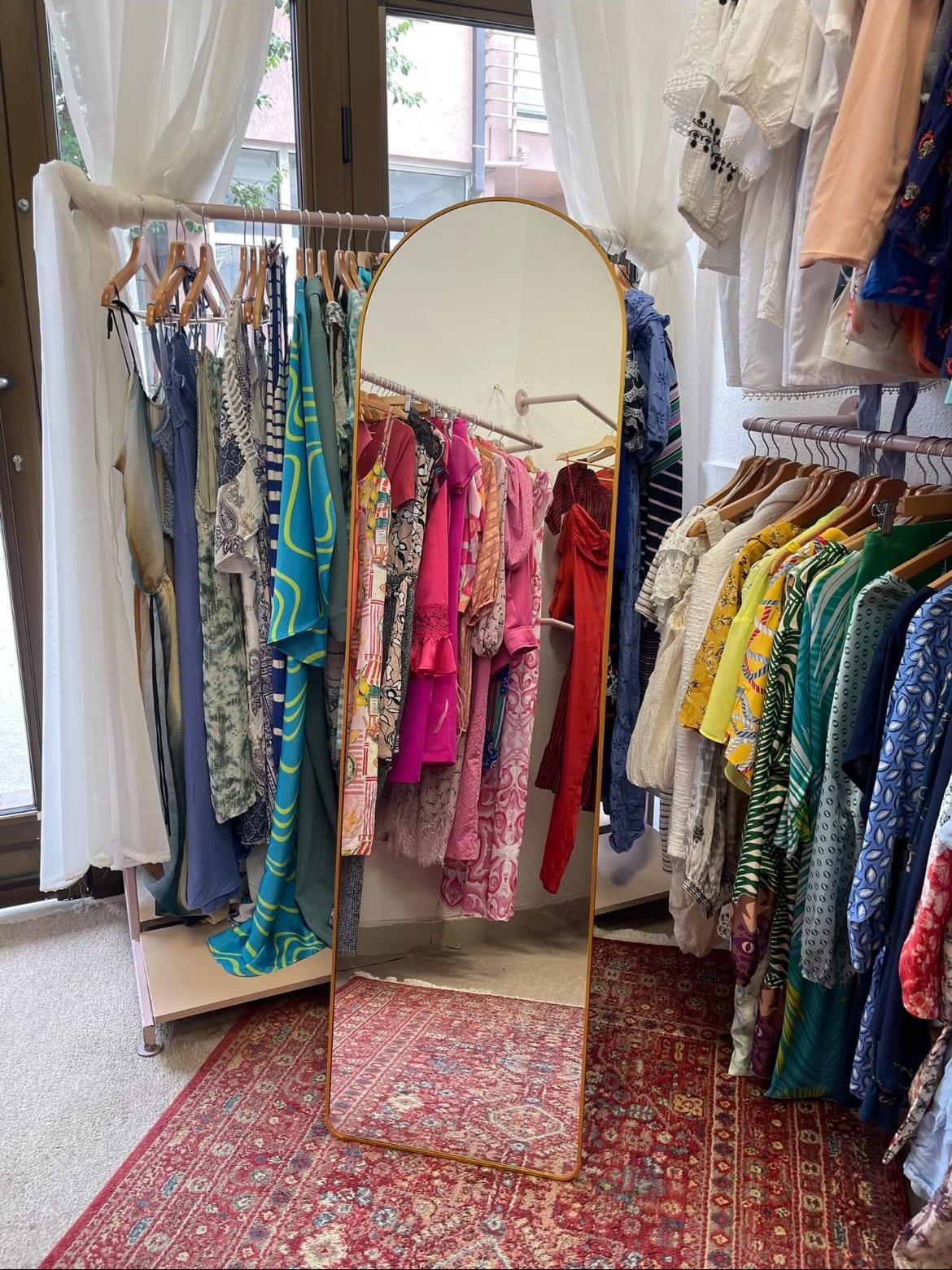 A gold arch mirror beside racks of clothing on a patterned rug inside the galerija boutique.